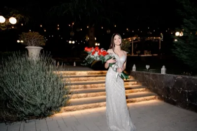 Ragazza in abito elegante argentato su una scalinata esterna illuminata di notte, con bouquet di rose rosse in primo piano. Sullo sfondo si vedono il giardino scuro, le luci calde appese tra gli alberi, grandi vasi decorativi e l'atmosfera scenografica dell'ingresso.