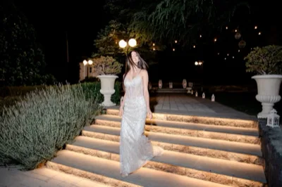 Ragazza sorridente in abito da sera argentato sulla scalinata del giardino di notte, ripresa in posa frontale con i capelli sciolti. Intorno ci sono gradini illuminati dal basso, piante decorative, lanterne e luci sospese che disegnano l'area esterna.