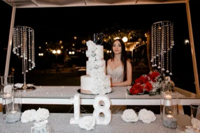 Ragazza in abito elegante accanto a un tavolo torta all'aperto di sera, sorridente mentre tiene un bouquet di rose rosse. Al centro si vede una torta bianca a più piani con decorazioni floreali e topper 18, affiancata da composizioni di rose bianche, candele e dettagli luminosi sotto una struttura coperta.