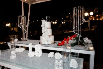 Tavolo scenografico per diciottesimo con torta bianca a più piani decorata da grandi rose bianche e numero 18 in cima. Sul tavolo ci sono candele, fiori rossi e bianchi, alzate trasparenti con dettagli brillanti e una cornice di luci calde sullo sfondo notturno.