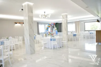 Sala luminosa con tavoli rotondi apparecchiati con tovaglie bianche, sedie bianche e runner azzurri, ripresa in una prospettiva ampia e ordinata. Al centro della sala c'è un arco di palloncini colorati, con tavolo decorativo sul fondo, grandi finestre, pilastri chiari e pavimento lucido che riflette la luce.