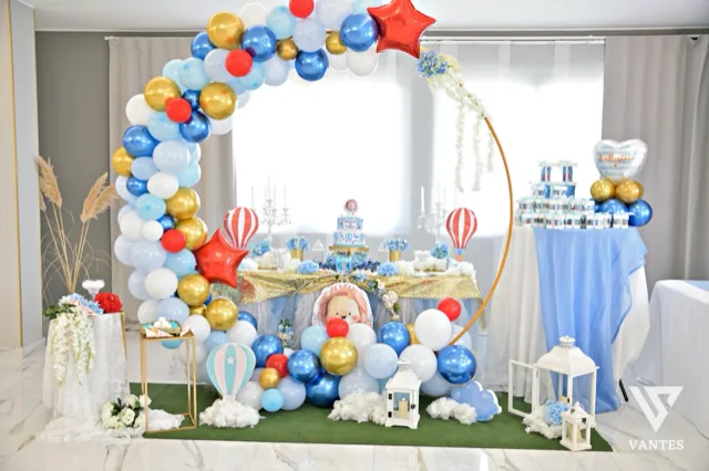 Allestimento scenografico per battesimo all'esterno o in sala, con grande arco di palloncini azzurri, bianchi, oro e rossi che incornicia il tavolo centrale. Sullo sfondo ci sono un tavolo dolci con confetti e bomboniere, elementi decorativi a tema baby, un palloncino a forma di cuore e grandi vasi floreali ai lati.