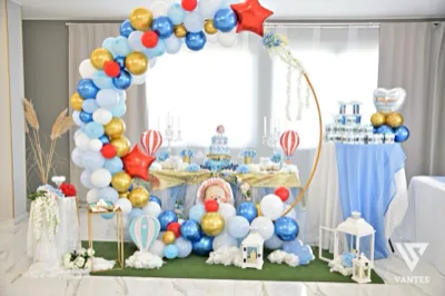 Allestimento scenografico per battesimo all'esterno o in sala, con grande arco di palloncini azzurri, bianchi, oro e rossi che incornicia il tavolo centrale. Sullo sfondo ci sono un tavolo dolci con confetti e bomboniere, elementi decorativi a tema baby, un palloncino a forma di cuore e grandi vasi floreali ai lati.