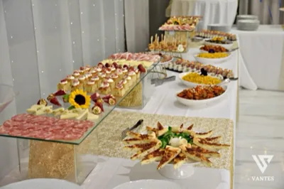 Buffet salato disposto su più livelli lungo un tavolo bianco con runner dorato, con taglieri di salumi, formaggi, stuzzichini, rustici, antipasti monoporzione e piatti caldi in file ordinate. Sullo sfondo si vedono piatti impilati, un fondale chiaro con lucine e una presentazione curata da catering per evento.