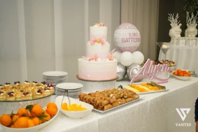 Tavolo dolci per battesimo con torta alta a tre piani in sfumature rosa e bianco, decorata con fiori delicati e piccoli frutti. Intorno ci sono monoporzioni, crostate, macedonia, piatti di dessert, arance fresche e un pallone decorativo con la scritta del battesimo sullo sfondo.