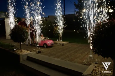 Momento scenografico notturno all'esterno con una bambina seduta su una piccola auto rosa, circondata da fontane a freddo che sparano scintille in alto. Accanto ci sono adulti che osservano la scena, il vialetto in pietra, cespugli illuminati e il cielo scuro del giardino.