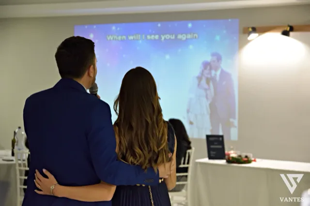 Due persone di spalle osservano una proiezione su grande schermo durante una festa in sala, mentre uno di loro tiene il microfono e l'immagine proiettata mostra una coppia in un momento romantico. Sullo sfondo si vedono tavoli apparecchiati, una parete chiara con applique e un'atmosfera intima da slideshow celebrativo.