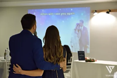 Due persone di spalle osservano una proiezione su grande schermo durante una festa in sala, mentre uno di loro tiene il microfono e l'immagine proiettata mostra una coppia in un momento romantico. Sullo sfondo si vedono tavoli apparecchiati, una parete chiara con applique e un'atmosfera intima da slideshow celebrativo.
