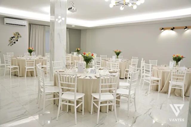 Sala interna con tavoli rotondi apparecchiati, tovaglie color sabbia, sedie bianche e piccoli centrotavola floreali su ogni tavolo. La scena mostra un allestimento ordinato e luminoso per il ricevimento, con pavimento chiaro effetto marmo e pareti neutre.