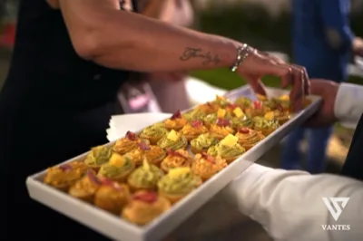 Vassoio rettangolare bianco con molti piccoli assaggi salati serviti a mano durante l'evento, disposti in file ordinate. Ogni boccone è decorato con creme, erbe e cubetti di formaggio, mentre le mani di chi serve e di chi riceve il vassoio occupano il primo piano.
