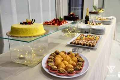 Buffet dei dolci allestito su un lungo tavolo con torte, frutta fresca, pasticcini e vassoi di dessert disposti su più livelli. In primo piano si vedono ciambelle glassate e piccoli dolci al cioccolato, mentre sul fondo ci sono piatti impilati, una bottiglia di spumante e altri vassoi da servizio.