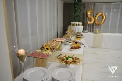 Tavolo buffet e torta per un cinquantesimo compleanno in sala, con grandi palloncini oro '50' sul fondo. In primo piano ci sono vassoi di antipasti, bottiglie di acqua e bibite, piatti impilati, mini portate e un ricco allestimento dolce e salato disposto su più livelli.