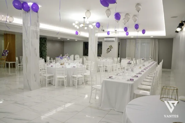Sala interna luminosa con tavoli rotondi e una lunga tavolata apparecchiata, sedie bianche e piccoli centrotavola viola. Dal soffitto scendono palloncini viola e bianchi, mentre i pilastri effetto marmo e il pavimento lucido danno un aspetto elegante e ordinato all'allestimento.