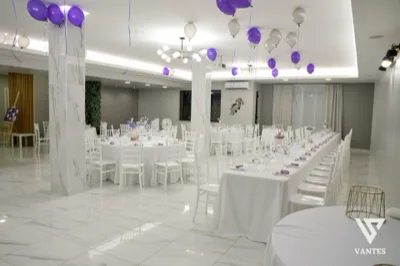 Sala interna luminosa con tavoli rotondi e una lunga tavolata apparecchiata, sedie bianche e piccoli centrotavola viola. Dal soffitto scendono palloncini viola e bianchi, mentre i pilastri effetto marmo e il pavimento lucido danno un aspetto elegante e ordinato all'allestimento.