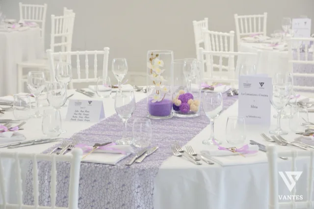 Primo piano di un tavolo rotondo apparecchiato con tovaglia bianca e runner lilla, bicchieri trasparenti, posate ordinate e segnaposto dedicati alla Comunione e Cresima. Al centro ci sono due cilindri decorativi con fiori bianchi e sfere lilla, mentre sullo sfondo si vedono altre tavole coordinate nella stessa palette.