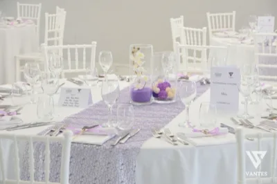 Primo piano di un tavolo rotondo apparecchiato con tovaglia bianca e runner lilla, bicchieri trasparenti, posate ordinate e segnaposto dedicati alla Comunione e Cresima. Al centro ci sono due cilindri decorativi con fiori bianchi e sfere lilla, mentre sullo sfondo si vedono altre tavole coordinate nella stessa palette.