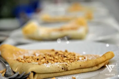 Primo piano di una crêpe dolce piegata e spolverata di zucchero a velo, con granella di frutta secca al centro e un'altra porzione sfocata sullo sfondo. L'immagine mette in evidenza la consistenza morbida del dessert e il servizio da buffet del dolce.