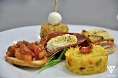 Primo piano di un piatto bianco con quattro assaggi salati curati nel dettaglio: una tartina con verdure, una crema decorata al centro, un piccolo sformato sul fondo e una porzione gialla compatta sulla destra. Il piatto è completato da foglie verdi e da una presentazione da buffet elegante e ravvicinata.