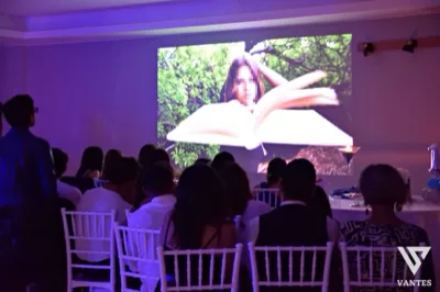Proiezione video in sala con il pubblico seduto su sedie bianche mentre sul grande schermo scorrono immagini della festeggiata all'aperto. La stanza è illuminata da luci viola e blu, con tavoli laterali apparecchiati e un'atmosfera raccolta da momento celebrativo.