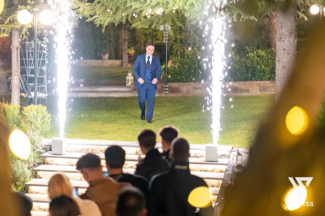 Ingresso scenografico notturno sul prato con due fontane luminose ai lati e un ragazzo in abito blu che avanza al centro dei gradini illuminati. In primo piano si vedono gli ospiti seduti e le luci calde del giardino, mentre il resto della scena è incorniciato da alberi e siepi curate.