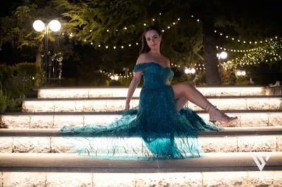 Festeggiata in abito lungo turchese seduta sulla scalinata esterna illuminata di notte, con il vestito che si apre ampio sui gradini. Sullo sfondo si vedono le luci calde del giardino, gli alberi e i lampioni che rendono la scena molto scenografica.