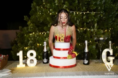 Tavolo torta all'aperto di notte con festeggiata in abito rosso e oro che soffia sulle candeline davanti a una torta a tre piani bianca e rossa. Sul tavolo ci sono numeri luminosi 18, bottiglie di spumante, un bicchiere cocktail a coppa e decorazioni floreali rosse e gialle su sfondo di siepe illuminata.