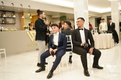 Momento in sala con un ragazzo che parla al microfono vicino al bancone e due giovani seduti in primo piano con abiti eleganti da cerimonia. Sullo sfondo ci sono altri ospiti, tavoli rotondi con tovaglie chiare e il banco bar illuminato, in un'atmosfera formale da ricevimento.