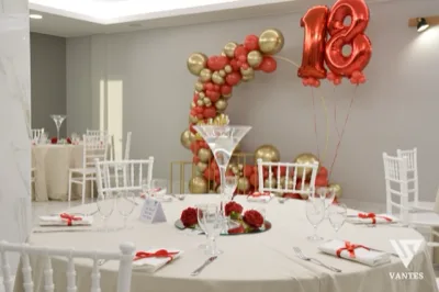 Tavolo rotondo apparecchiato con tovaglia chiara, calici, tovaglioli con nastri rossi e centrotavola floreale rosso al centro. Sullo sfondo si vede un arco di palloncini rosso e oro con numero 18 metallico, insieme ad altri tavoli coordinati nella sala.