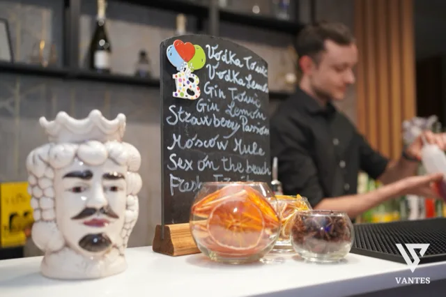 Angolo bar in primo piano con lavagna nera scritta a mano con nomi di cocktail, ciotole di vetro con fettine di agrumi e una maschera decorativa bianca a lato. Sullo sfondo si vede il barman al lavoro dietro il bancone, in una scena calda e molto ravvicinata del servizio drink.