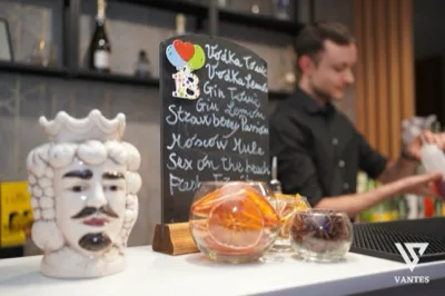 Angolo bar in primo piano con lavagna nera scritta a mano con nomi di cocktail, ciotole di vetro con fettine di agrumi e una maschera decorativa bianca a lato. Sullo sfondo si vede il barman al lavoro dietro il bancone, in una scena calda e molto ravvicinata del servizio drink.
