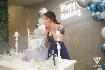 Festeggiata in abito blu scuro mentre soffia sulle candeline davanti a una torta a due piani decorata con fiori lilla e azzurri. Sullo sfondo ci sono palloncini blu e argento, la scritta luminosa Happy Birthday e piccoli dettagli da tavolo torta con palette coordinata.