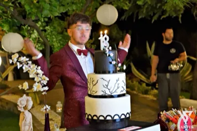 Ragazzo in giacca bordeaux accanto a una torta a tre piani bianca e nera con candeline a forma di 18, durante un festeggiamento serale in giardino. Sullo sfondo si vedono lanterne di carta appese agli alberi, piante ornamentali e un addetto di sala in un'atmosfera raccolta da taglio torta.