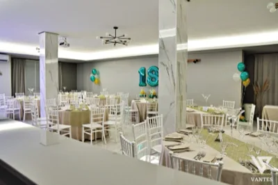 Sala interna apparecchiata per un diciottesimo compleanno con tavoli rotondi, sedie bianche e runner verde salvia su tovaglie color avorio. Sullo sfondo si vedono palloncini numerici 18 in verde petrolio, piccoli gruppi di palloncini coordinati e un ambiente luminoso con pilastri effetto marmo.