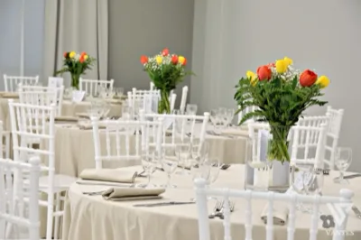 Dettaglio di una sala interna apparecchiata con tavoli rotondi, sedie bianche e tovagliato avorio in un ambiente luminoso e minimale. In primo piano spicca un centrotavola con tulipani rossi e gialli, ripetuto anche sugli altri tavoli insieme a calici ordinati e segnaposto discreti.