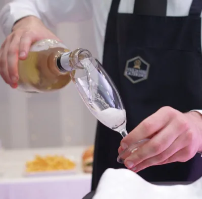 Primo piano di un cameriere che versa spumante in un flute durante il servizio, con il flusso del vino e la schiuma ben visibili. Sullo sfondo si intravede un tavolo del buffet sfocato, in una scena elegante e ravvicinata di accoglienza al ricevimento.