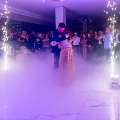 Coppia che si abbraccia al centro della pista da ballo mentre le fontane luminose sparano scintille ai lati e una nube di fumo scenico avvolge il pavimento. Gli ospiti attorno osservano e riprendono il momento con i telefoni, sotto una luce viola intensa da serata evento.
