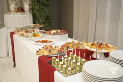 Buffet salato allestito su una lunga tavola bianca con runner bordeaux, piatti impilati e numerosi assaggi monoporzione disposti su alzate e vassoi. In primo piano si vedono cubi di torta salata, tramezzini e piccoli stuzzichini, mentre sullo sfondo ci sono piatti di antipasti, salumi e portate da aperitivo elegante.