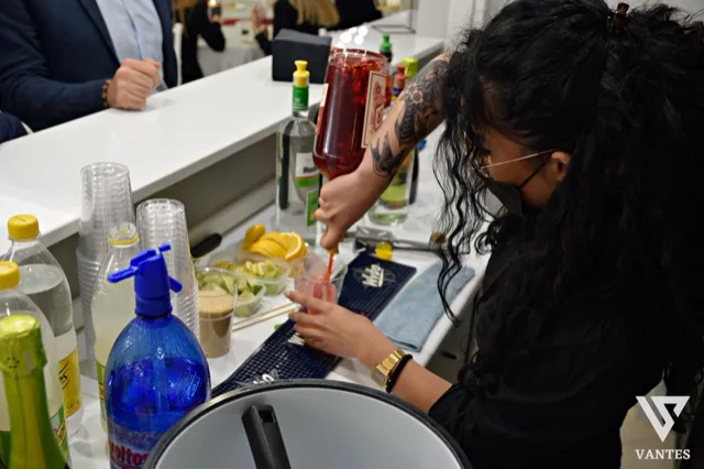 Barista al banco mentre prepara un cocktail rosso versandolo in un bicchiere con ghiaccio, con bottiglie di liquori, bicchieri impilati, bottigliette d'acqua e frutta tagliata disposti sul piano. La scena è ravvicinata e mostra il servizio drink durante la festa di laurea.