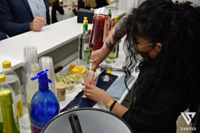 Barista al banco mentre prepara un cocktail rosso versandolo in un bicchiere con ghiaccio, con bottiglie di liquori, bicchieri impilati, bottigliette d'acqua e frutta tagliata disposti sul piano. La scena è ravvicinata e mostra il servizio drink durante la festa di laurea.