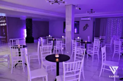 Ampia lounge interna con luce viola diffusa, tavolini rotondi neri, sedie bianche, pouf quadrati e piccole luci decorative sul fondo. L'ambiente è ordinato e minimale, pensato come area relax del ricevimento di laurea, con pavimento lucido e pareti chiare.