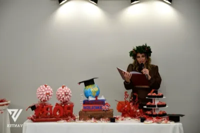 Tavolo torta per laurea con una giovane laureata che legge o parla al microfono davanti a un allestimento rosso e arancione. Sul tavolo ci sono dolci decorati, la scritta 'FRANCAIS ENGLISH' su un libro decorativo, un globo con tocco da laurea e grandi elementi con il nome della festeggiata.