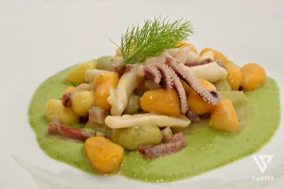 Primo piano di un piatto gourmet con crema verde alla base, cubetti di patate, pezzi di polpo, piccoli tagli di salume e aneto fresco sopra. La composizione è impiattata con cura su un piatto bianco ampio, in stile fine dining da ricevimento di matrimonio.