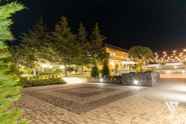 Ampia vista notturna del giardino con pavimentazione in mattoni, alberi alti illuminati dal basso, villa gialla sullo sfondo e catenarie di luci calde sospese sopra l'area esterna. La scena mostra lo spazio evento vuoto e scenografico, pronto per il ricevimento di matrimonio.