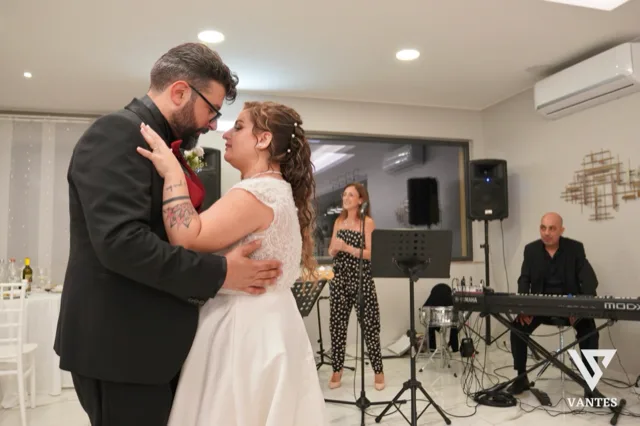 Gli sposi ballano abbracciati in sala durante il ricevimento, con lei in abito bianco e lui in completo scuro con dettaglio rosso. Sullo sfondo si vedono la cantante, il tastierista, i microfoni e le casse della band, in un momento intimo e musicale della festa.