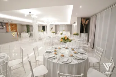Ampia sala interna molto luminosa con tavoli rotondi apparecchiati, tovaglie bianche, sedie bianche e centrotavola floreali giallo-arancio al centro di ogni tavolo. Il pavimento lucido effetto marmo, le tende morbide e il banco bar sul lato sinistro creano un allestimento elegante e minimale per il matrimonio.
