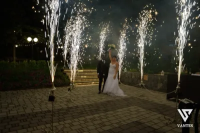 Gli sposi posano al centro del piazzale notturno circondati da alte fontane luminose che sparano scintille verso l'alto. Il marito è in abito scuro e la sposa in abito bianco solleva il bouquet, mentre sullo sfondo si vedono i gradini illuminati e le aiuole del giardino.
