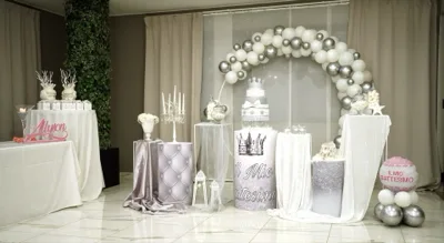 Allestimento in sala per battesimo con grande arco di palloncini bianchi e argento che incornicia il tavolo della torta sul fondo. La scenografia include cilindri decorativi rivestiti in tessuto e glitter, candelabro bianco, bomboniere coordinate e un pallone rosa con scritta dedicata al battesimo.