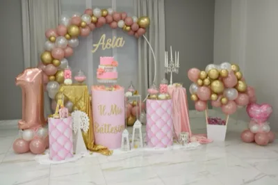 Scenografia in sala per battesimo e primo compleanno con arco di palloncini rosa, bianco e oro, torta a più piani al centro e grande palloncino a forma di 1 sulla sinistra. Intorno si vedono cilindri decorativi trapuntati, candelabro bianco, lanterne a terra, vasi con confetti e un grande mazzo di palloncini coordinati sulla destra.