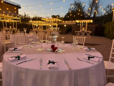 Allestimento esterno serale con tavoli rotondi apparecchiati, sedie bianche, candele e bicchieri disposti su una tovaglia chiara, sotto catenarie di luci calde sospese tra i pergolati. La scena mostra più aree conviviali illuminate al tramonto, con atmosfera elegante e rilassata.