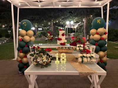 Allestimento serale all'aperto per un diciottesimo con tavolo bianco a più livelli, torta a piani al centro e numero 18 luminoso in primo piano. Ai lati ci sono colonne di palloncini verde petrolio e oro, mentre rose rosse, bomboniere e un runner dorato completano la scenografia sotto una struttura illuminata nel giardino.